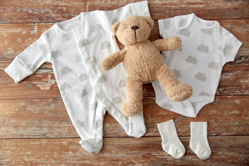 baby clothes, babyhood and clothing concept - bodysuits and socks with teddy bear toy on wooden table. baby bodysuits and teddy bear on wooden table