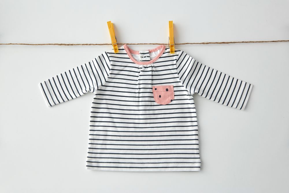 clothes, laundry, babyhood and clothing concept - striped long-sleeved shirt for baby girl with pink pocket hanging on clothesline with pins on white background. shirt for baby girl hanging on rope with pins