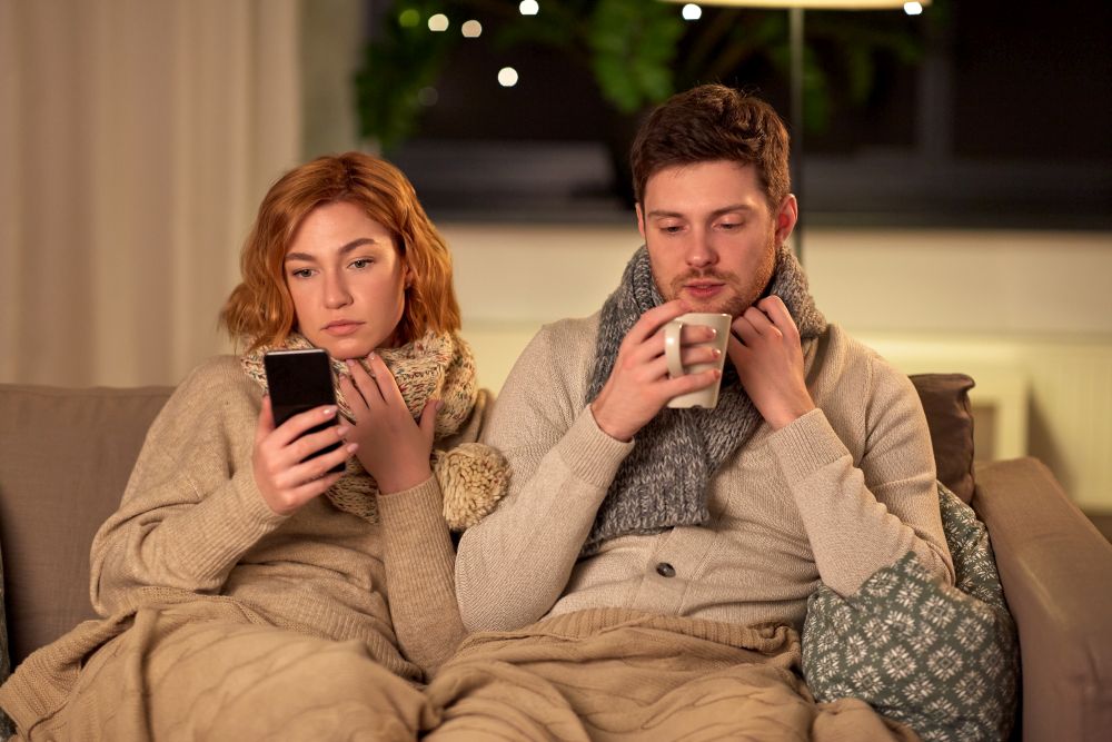 health, flu and people concept - sick young couple in scarves with smartphone drinking tea at home. sick young couple with smartphone at home