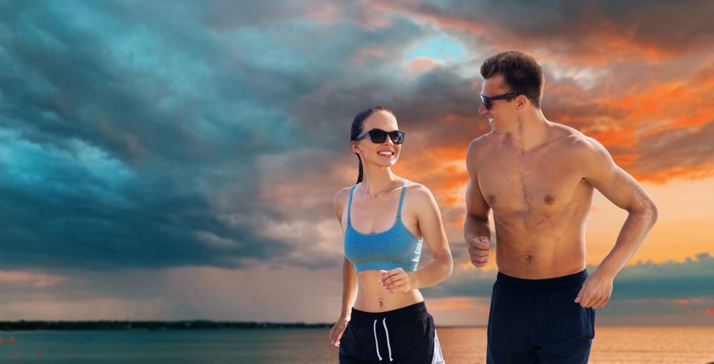 fitness, sport and lifestyle concept - happy couple in sports clothes and sunglasses running over sea and sunset sky on background. couple in sports clothes running along on beach