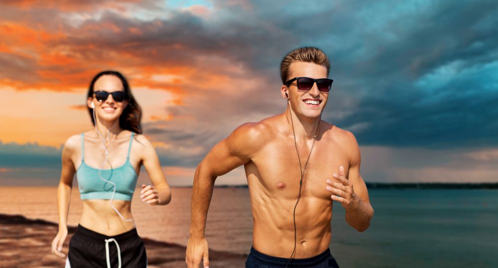 fitness, sport and technology concept - happy couple with earphones running over sea and sunset sky on background. couple with earphones running over sea