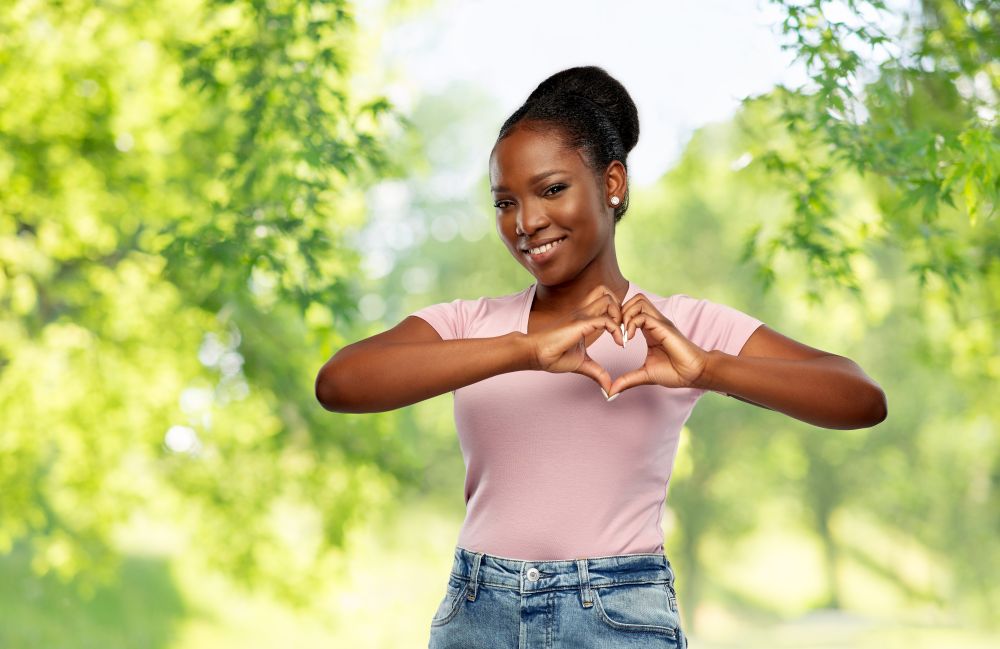 love, eco and valentine&rsquo;s day concept - happy smiling african american young woman making hand heart gesture over green natural background. african american woman making hand heart gesture