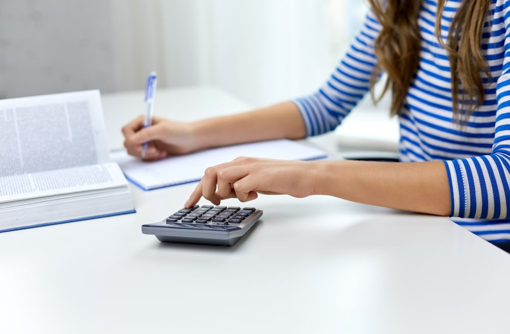 education, school and knowledge concept - close up of student girl counting on calculator and writing to notebook at home. student girl counting on calculator at home