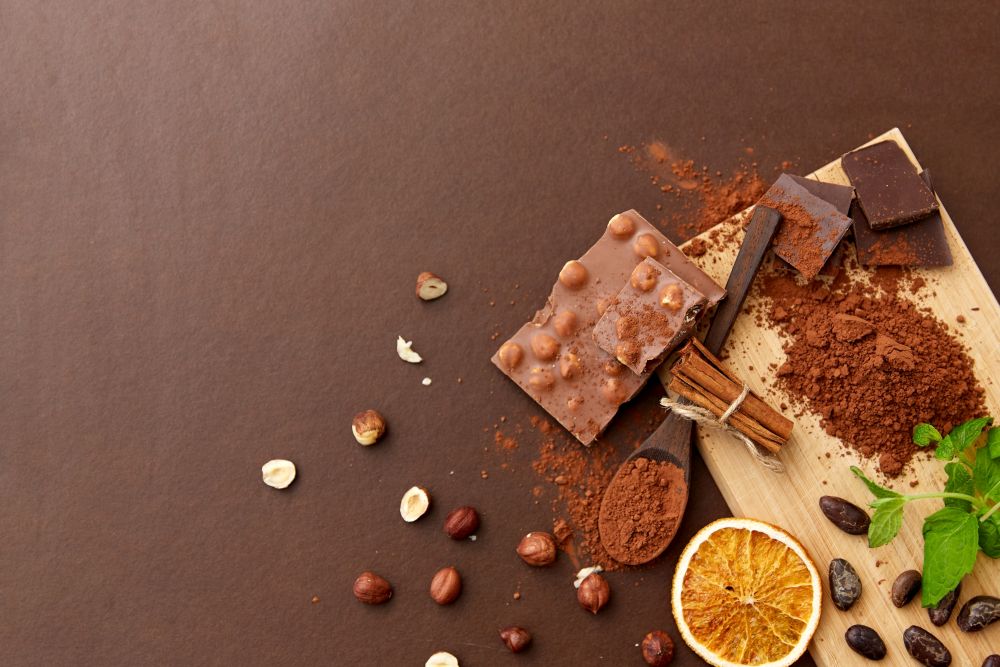 sweets, confectionery and culinary concept - chocolate with hazelnuts, cocoa beans, and powder with dry orange and cinnamon on wooden board with spoon on brown background. chocolate with hazelnuts, cocoa beans and powder