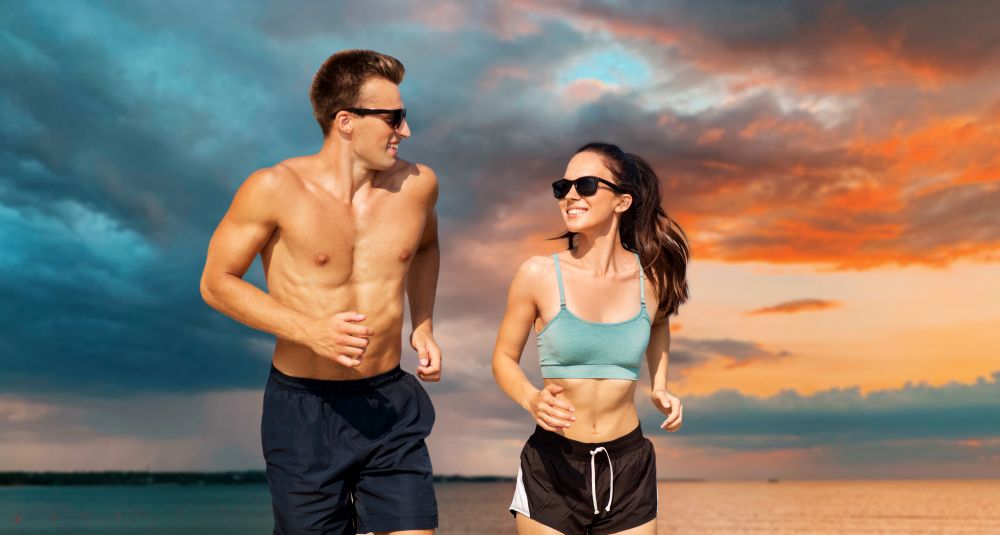 fitness, sport and lifestyle concept - happy couple in sports clothes and sunglasses running over sea and sunset sky on background. couple in sports clothes running along on beach