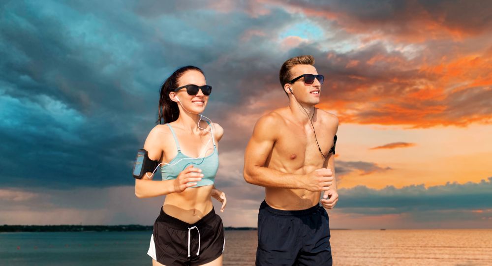 fitness, sport and technology concept - happy couple with earphones and arm bands running over sea and sunset sky on background. couple with phones and arm bands running over sea