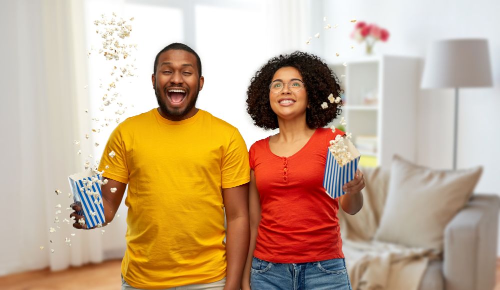 food, entertainment and people concept - happy smiling african american couple with popcorn flying in air over home background. happy african american couple with popcorn