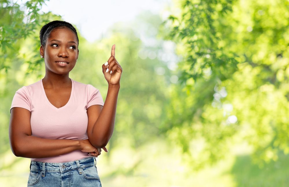 people, idea and attention concept - happy african american young woman pointing finger up over green natural background. happy african american woman pointing finger up