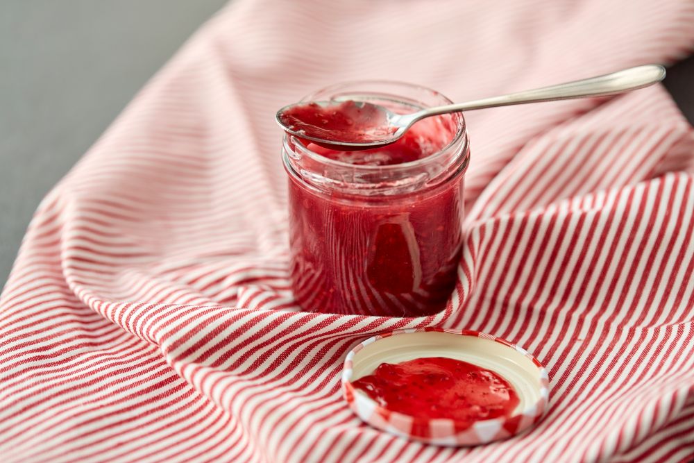 food, preserves and eating concept - mason jar with red raspberry jam and spoon on towel. mason jar with raspberry jam and spoon on towel