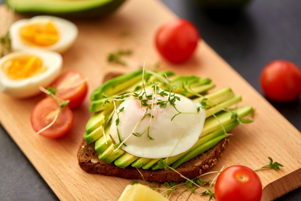 food, eating and breakfast concept - toast bread with sliced avocado, pouched egg, cherry tomatoes and greens on wooden cutting board. toast bread with avocado, eggs and cherry tomatoes