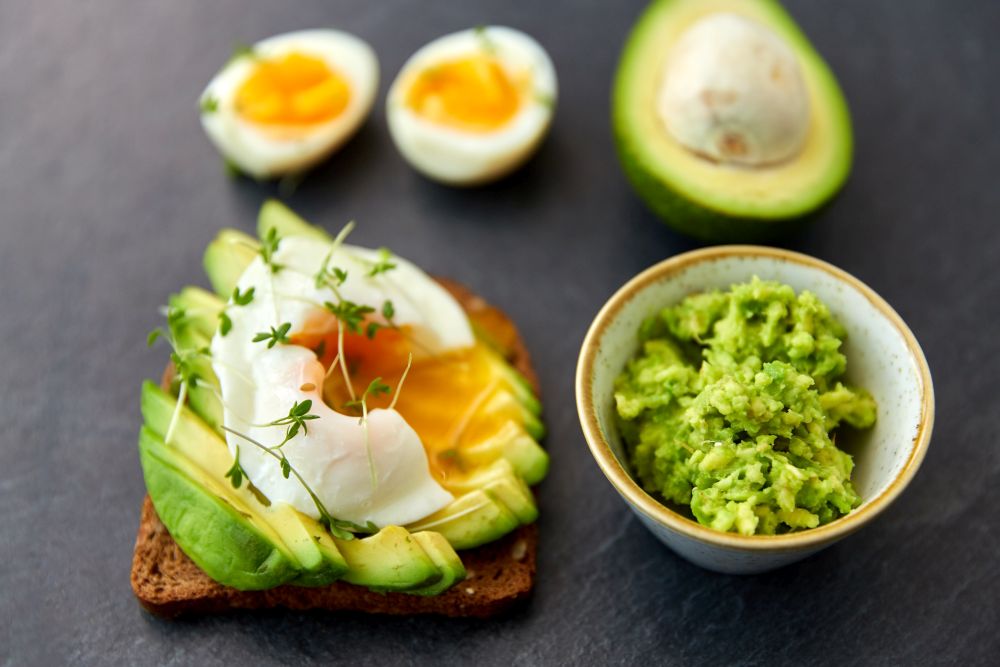 food, eating and breakfast concept - toast bread with sliced avocado, pouched egg and greens on slate stone table. toast bread with avocado, pouched egg and greens