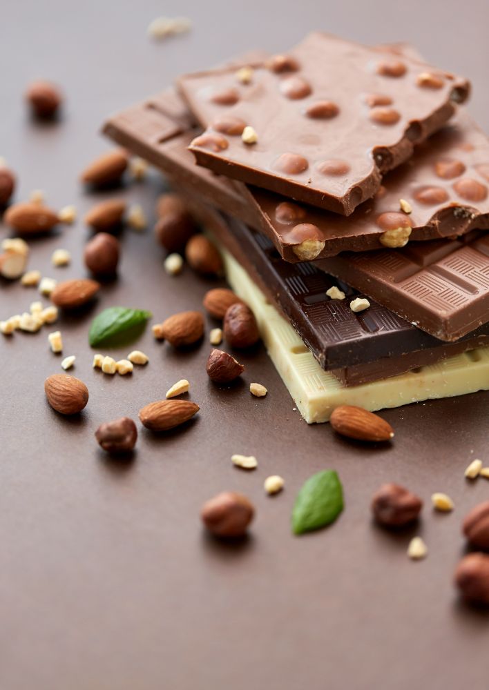 sweets, confectionery and food concept - bars of dark, white and milk chocolate with nuts on brown background. close up of different chocolate bars and nuts