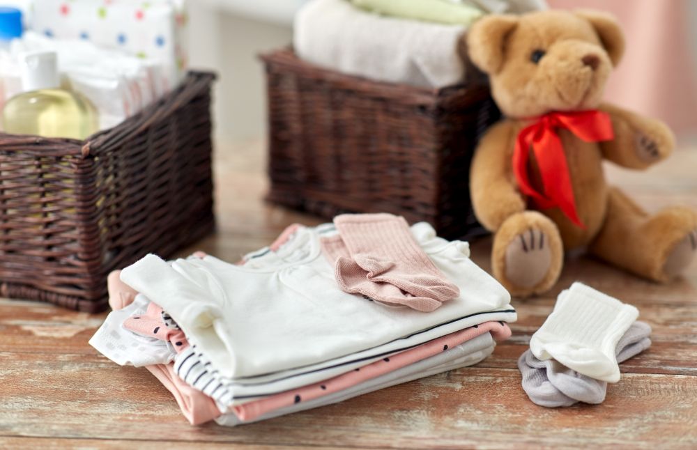 babyhood and clothing concept - baby clothes, teddy bear toy and baskets on wooden table at home. baby clothes and teddy bear toy on table at home