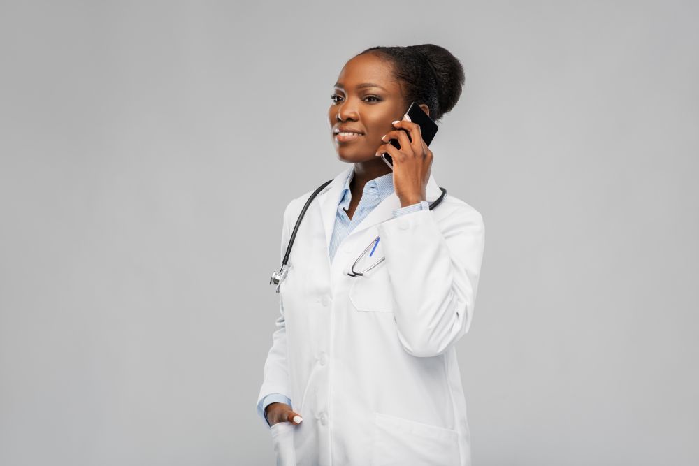medicine, technology and healthcare concept - happy smiling african american female doctor or in white coat with stethoscope calling on smartphone over background. african female doctor calling on smartphone