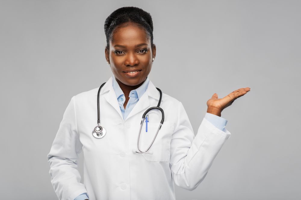 medicine, profession and healthcare concept - happy smiling african american female doctor in white coat with stethoscope showing something imnaginary over background. african american female doctor with stethoscope