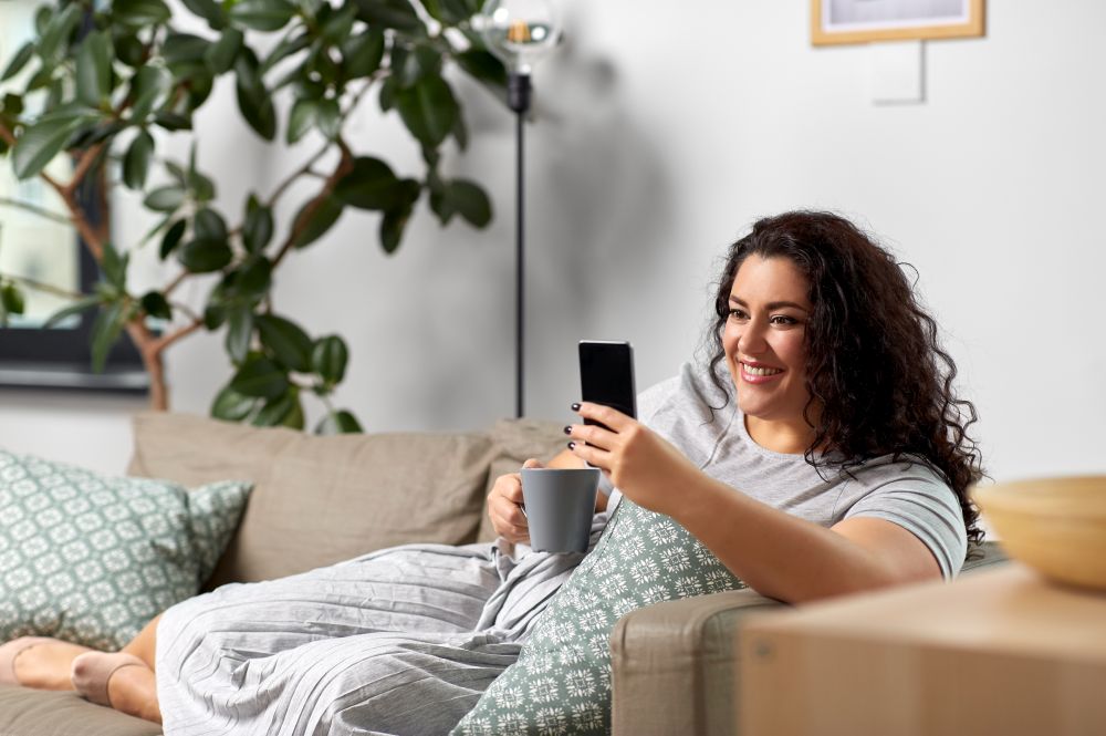 technology, leisure and people concept - happy smiling woman with smartphone drinking tea or coffee at home. woman with smartphone drinking coffee at home