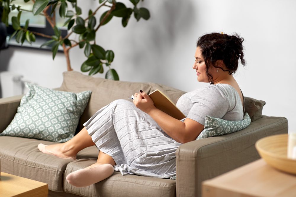 people and leisure concept - happy young woman with diary and pencil taking notes on sofa at home. happy young woman with diary on sofa at home