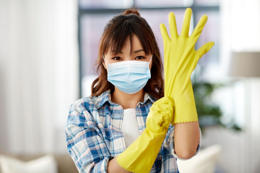 cleaning, health and hygiene concept - asian woman wearing protective medical mask for protection from virus disease and putting rubber gloves on at home. asian woman in protective mask and rubber gloves