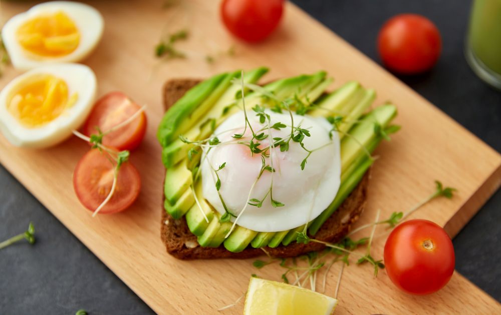 food, eating and breakfast concept - toast bread with sliced avocado, pouched egg, cherry tomatoes and greens on wooden cutting board. toast bread with avocado, eggs and cherry tomatoes