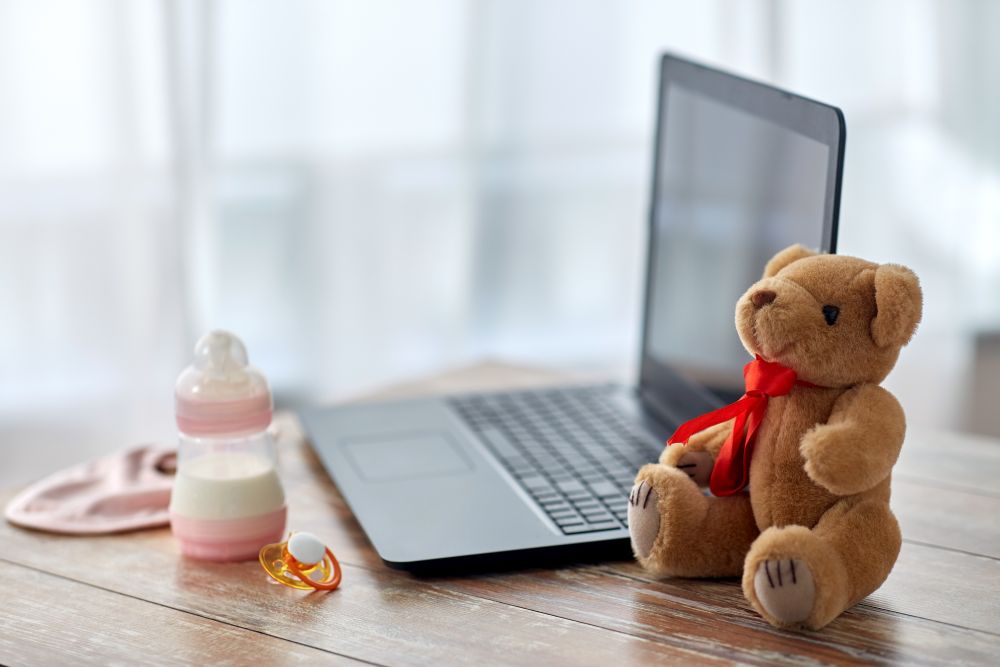 feeding and technology concept - bottle with baby milk formula, laptop computer, teddy bear toy and soother on wooden table at home. baby milk formula, laptop, soother and teddy bear