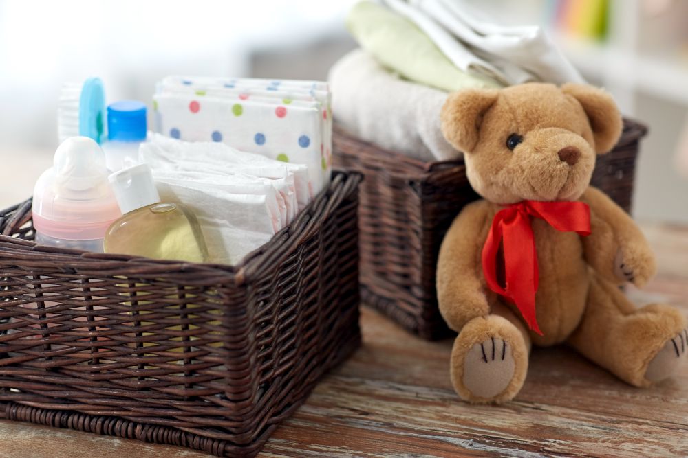 babyhood and care products concept - baby things in wicker baskets and teddy bear toy on wooden table at home. baby things in baskets and teddy bear toy on table
