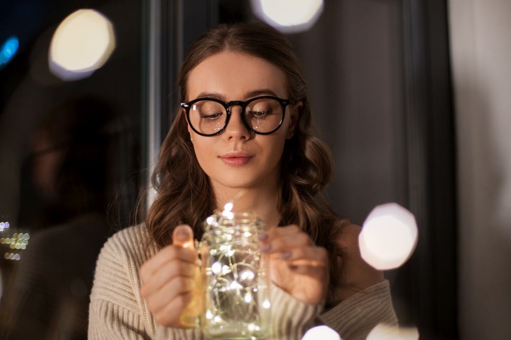 winter, comfort and people concept - young woman in glasses with garland lights in mason jar mug at home. woman with garland lights in glass mug at home