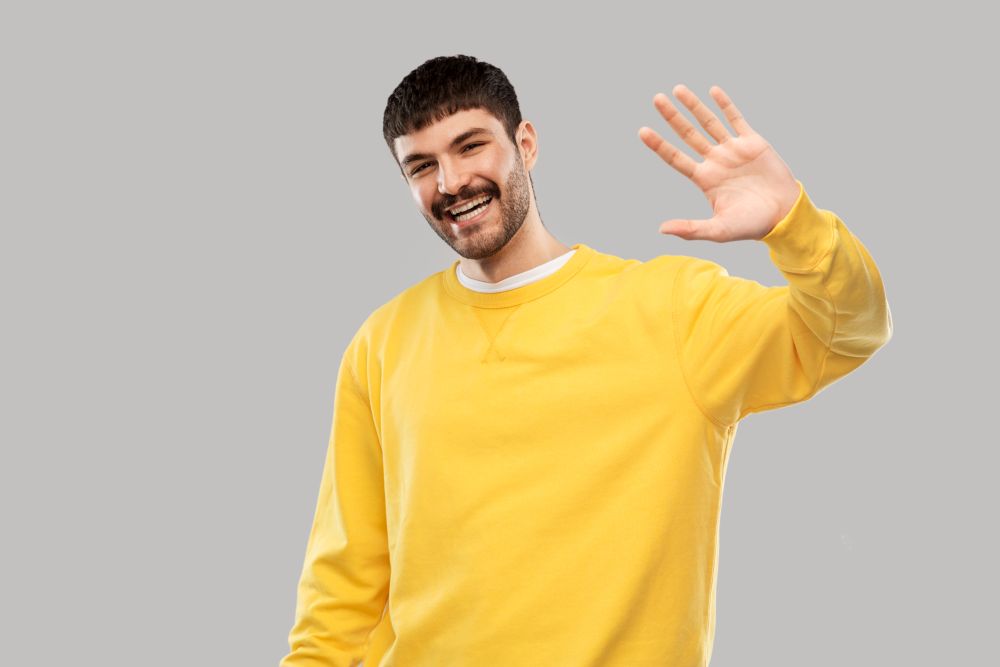 gesture and people concept - smiling young man in yellow sweatshirt waving hand over grey background. smiling young man in yellow sweatshirt waving hand
