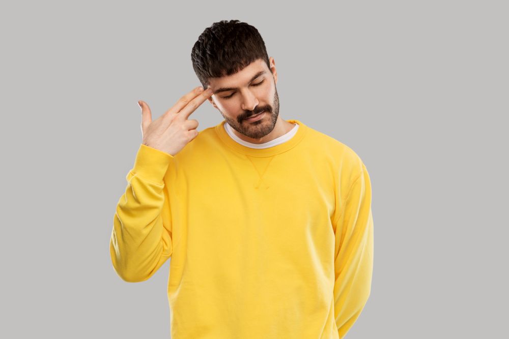 people, expression and stress concept - bored young man in yellow sweatshirt making headshot by finger gun gesture. bored man making finger gun gesture
