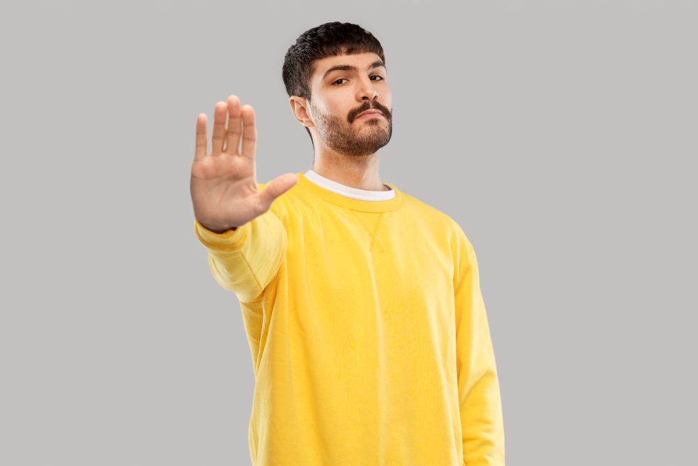 warning and people concept - serious young man in yellow sweatshirt showing stop gesture over grey background. serious young man showing stop gesture