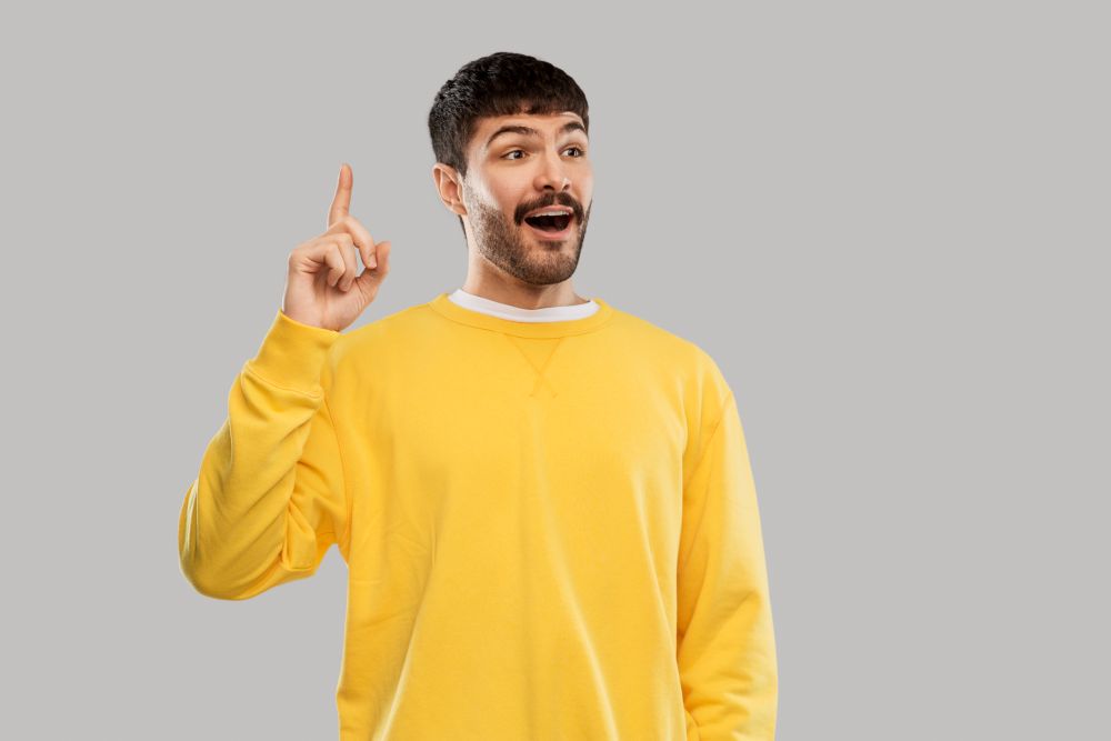 count and people concept - smiling young man in yellow sweatshirt showing one finger over grey background. young man showing one finger in yellow sweatshirt