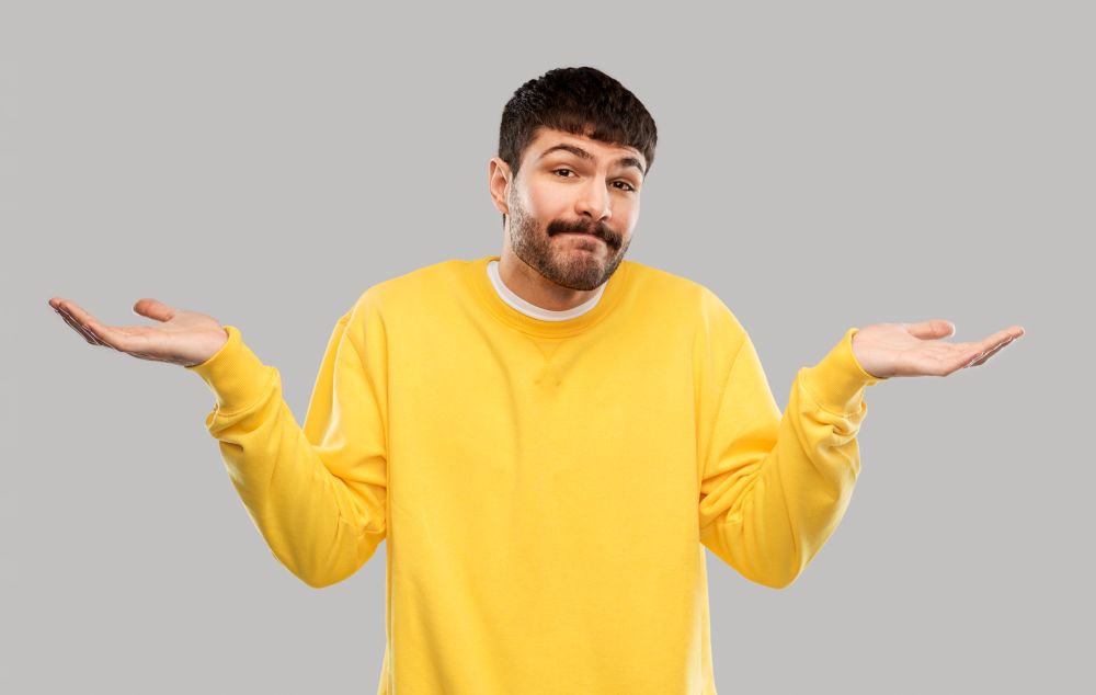 people, emotion and expression concept - confused man in yellow sweatshirt shrugging over grey background. confused man in yellow sweatshirt shrugging