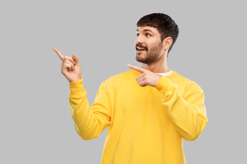 people concept - smiling young man in yellow sweatshirt pointing fingers to something over grey background. smiling man pointing fingers to something