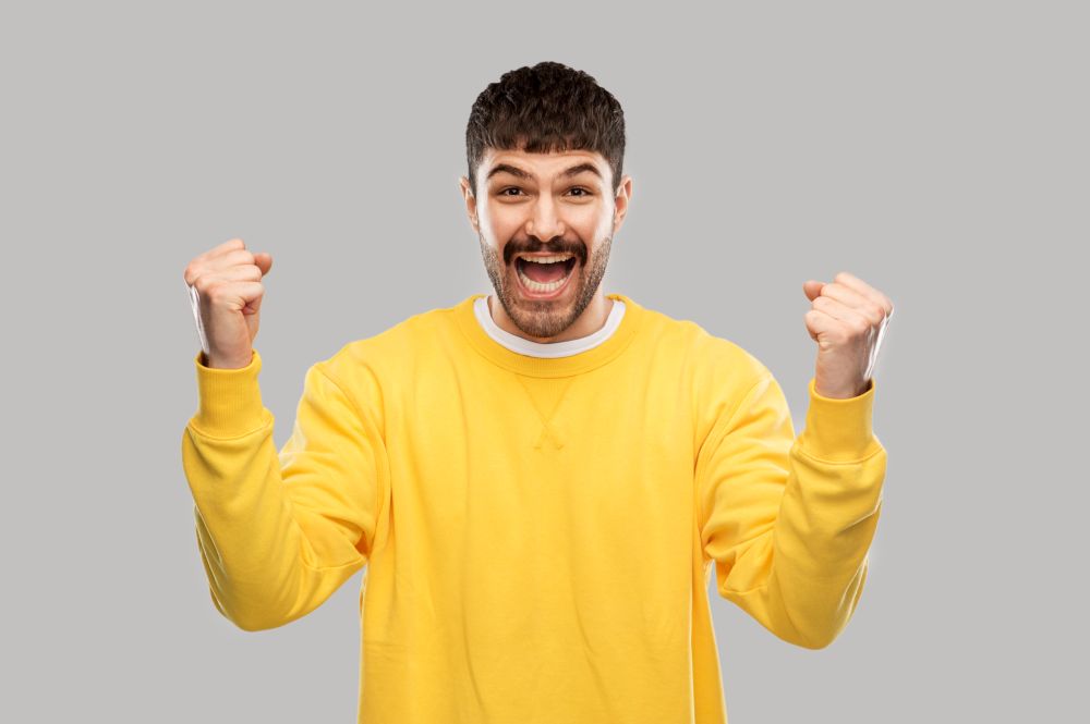 success, emotion and expression concept - happy man in yellow sweatshirt celebrating victory over grey background. happy man in yellow sweatshirt celebrating victory