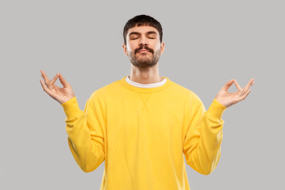 yoga, meditation and mindfulness concept - young man in yellow sweatshirt meditating over grey background. young man in yellow sweatshirt meditating