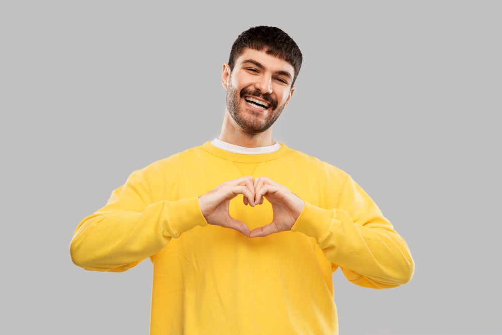 love, valentine&rsquo;s day and people concept - smiling young man in yellow sweatshirt girl making hand heart gesture over grey background. smiling young man in yellow sweatshirt