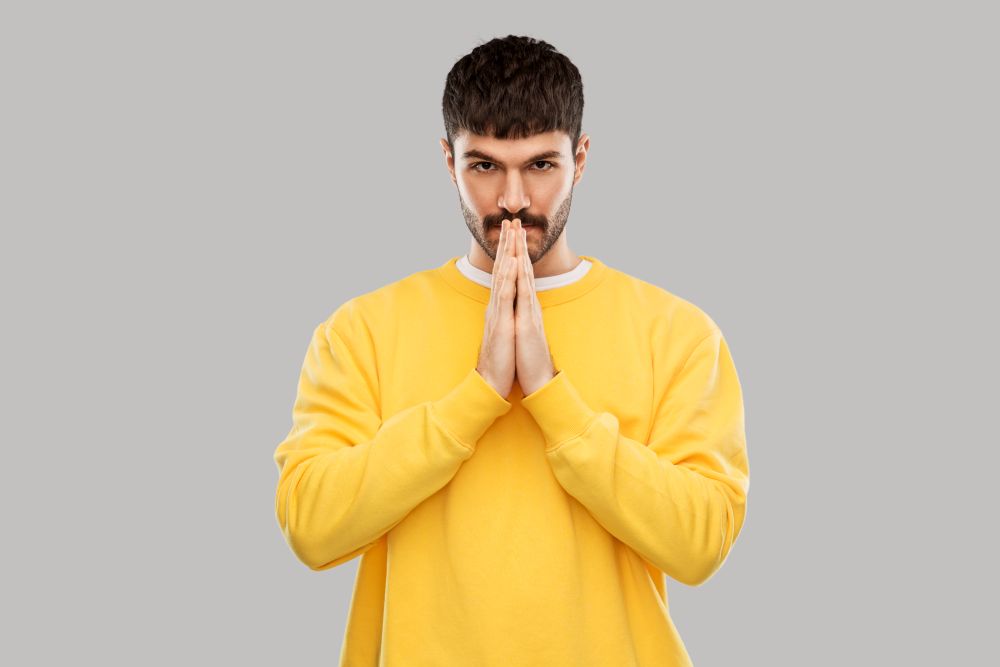 meditation, faith and mindfulness concept - young man in yellow sweatshirt praying or giving thanks over grey background. man in yellow sweatshirt praying or giving thanks