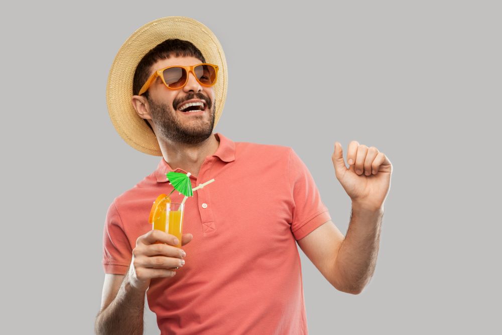 summer, vacation and people concept - happy smiling young man in sunglasses and straw hat with orange juice cocktail dancing over grey background. happy man in straw hat with orange juice cocktail