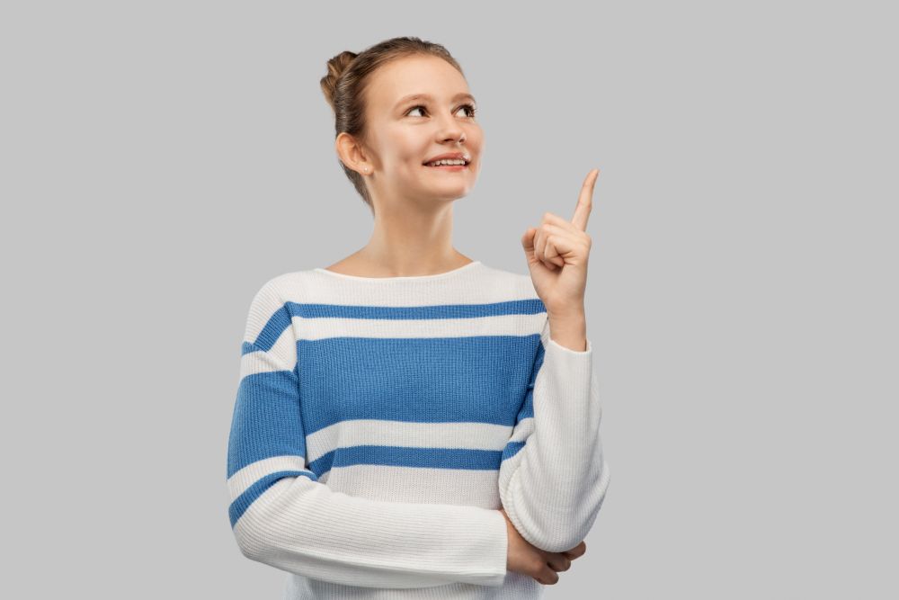idea and people concept - smiling teenage girl in pullover pointing finger up over grey background. happy teenage girl in pullover pointing finger up