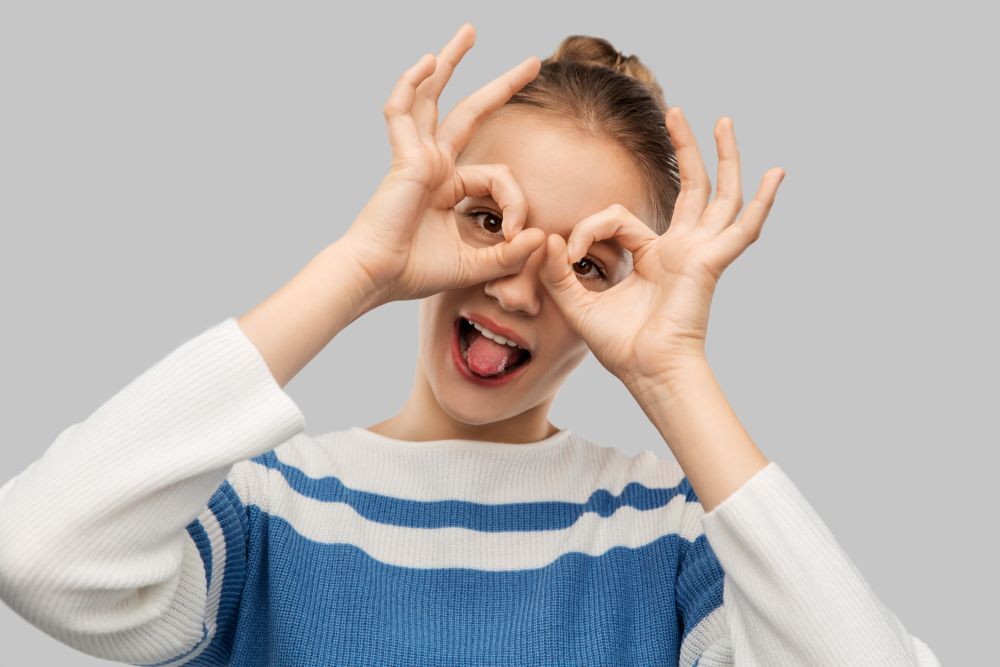 people concept - funny teenage girl looking through finger glasses over grey background. funny teenage girl looking through finger glasses