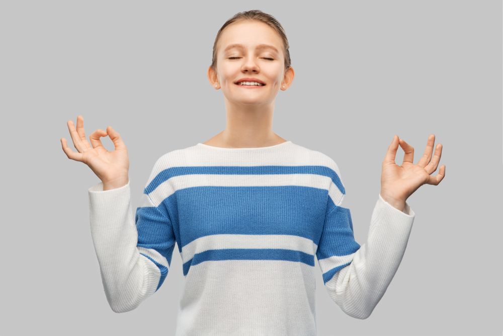 meditation and mindfulness people concept - happy smiling teenage girl in pullover meditating over grey background. happy smiling teenage girl in pullover meditating
