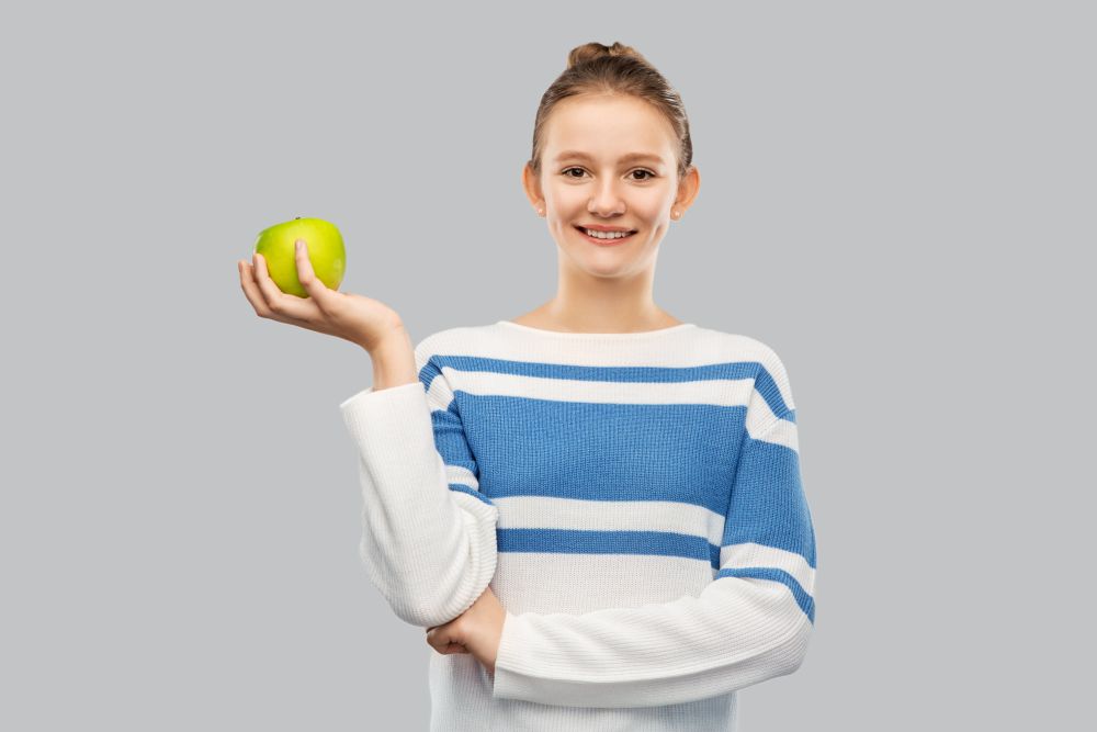 healthy eating, food and people concept - happy smiling teenage girl in pullover holding green apple over grey background. smiling teenage girl in pullover with green apple