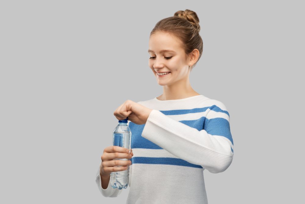 drink, health and people concept - happy smiling teenage girl in pullover opening bottle of water over grey background. smiling teenage girl opening bottle of water