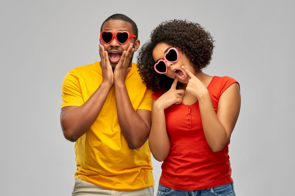 love, relationships and valentines day concept - happy smiling african american couple in heart shaped sunglasses making silly faces over grey background. happy african couple in heart shaped sunglasses