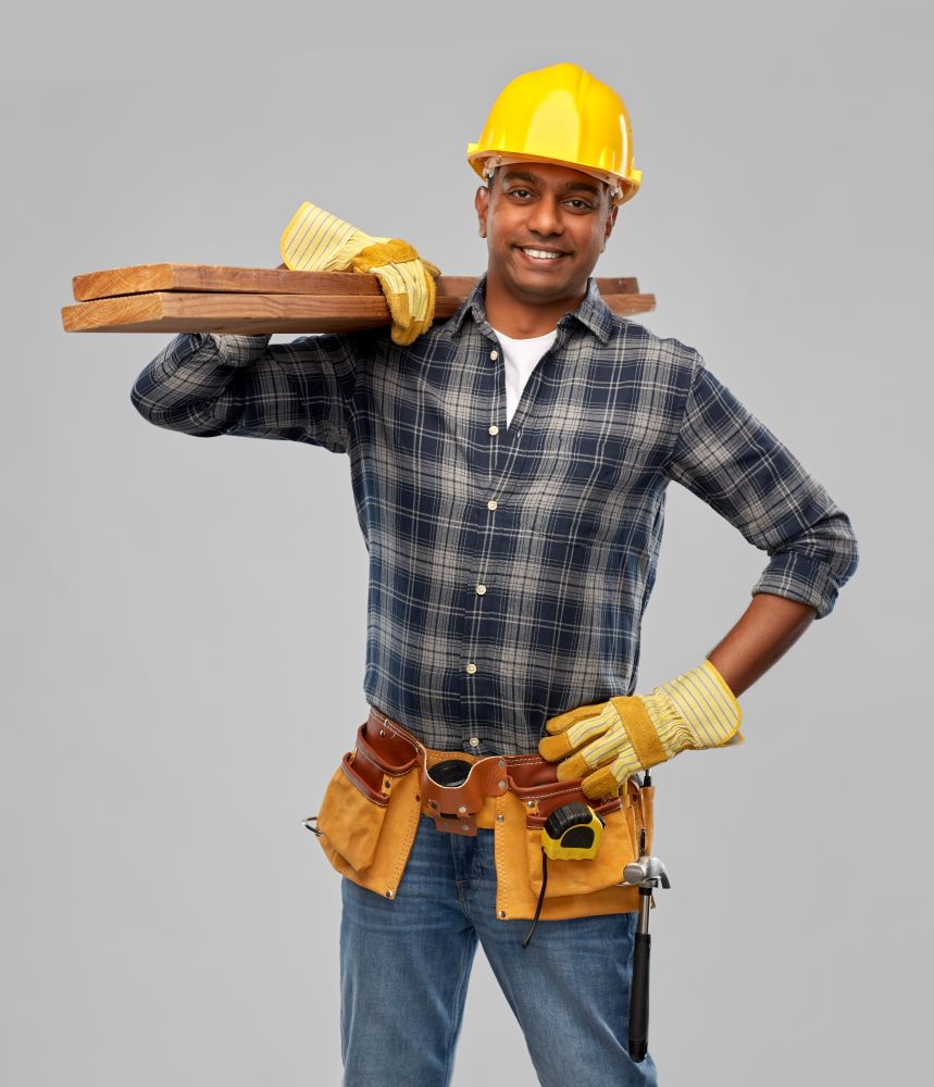 profession, construction and building - happy smiling indian carpenter or builder in helmet with tool belt and wooden boards over grey background. happy indian worker or builder with wooden boards