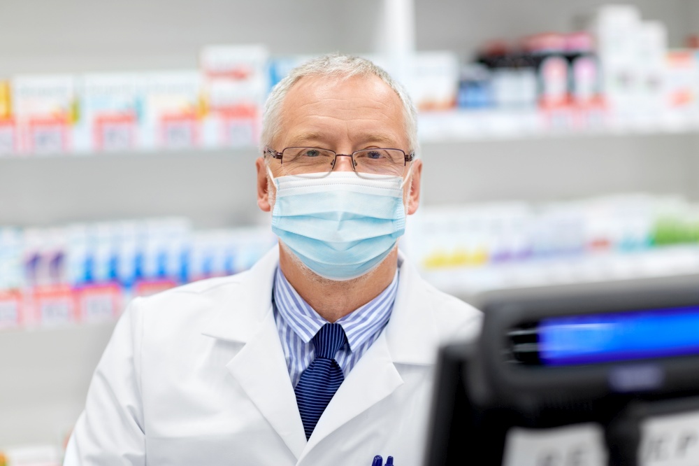 medicine, healthcare and people concept - senior apothecary wearing face protective medical mask for protection from virus disease at pharmacy cash register. senior apothecary in mask at pharmacy