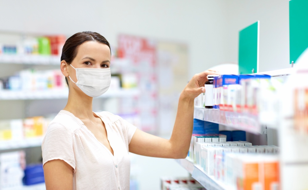 medicine, healthcare and people concept - female customer in face protective medical mask for protection from virus disease choosing drugs at pharmacy. female customer in mask choosing drugs at pharmacy