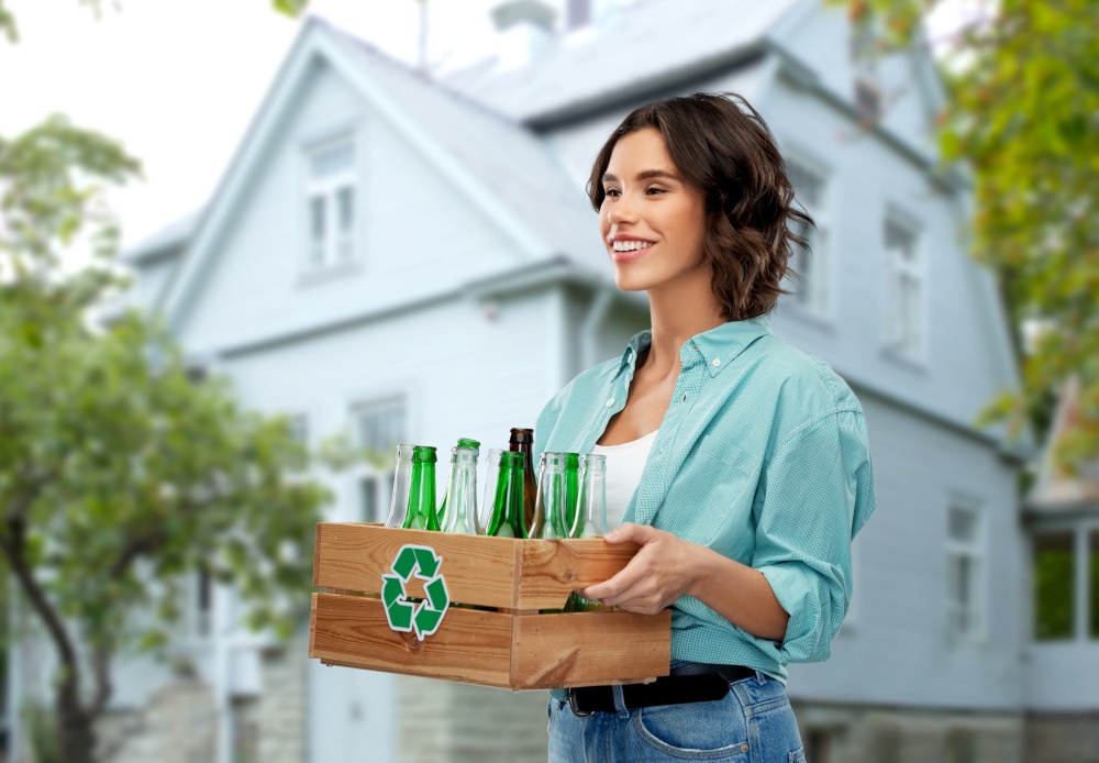 recycling, waste sorting and sustainability concept - smiling young woman holding wooden box with glass bottles and jars over house background. smiling young woman sorting glass waste outdoors
