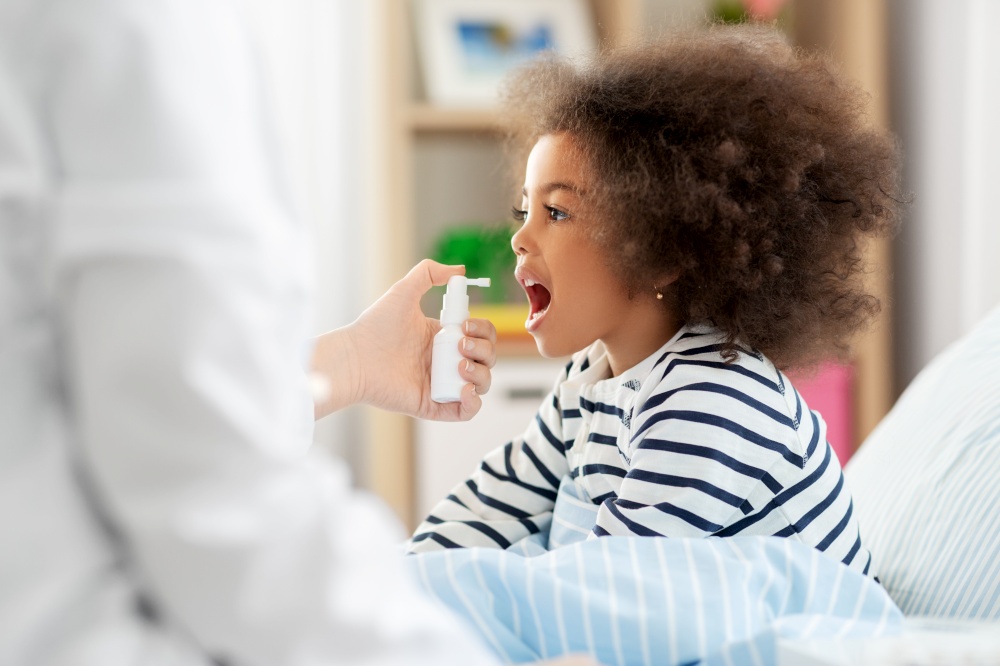 healthcare, medicine and people concept - doctor with oral spray treats little sick african american girl in bed at home. doctor with medicine treats sick girl at home
