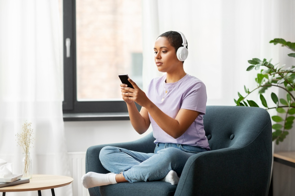 people, technology and leisure concept - young african american woman in glasses with smartphone and headphones sitting in chair and listening to music at home. woman with smartphone listening to music at home