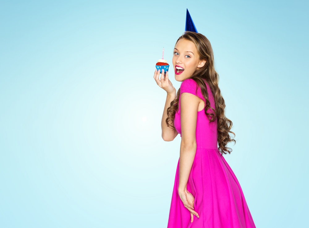 people and celebration concept - happy young woman or teen girl in pink dress and party cap with birthday cupcake over blue background. happy woman or teen girl with birthday cupcake